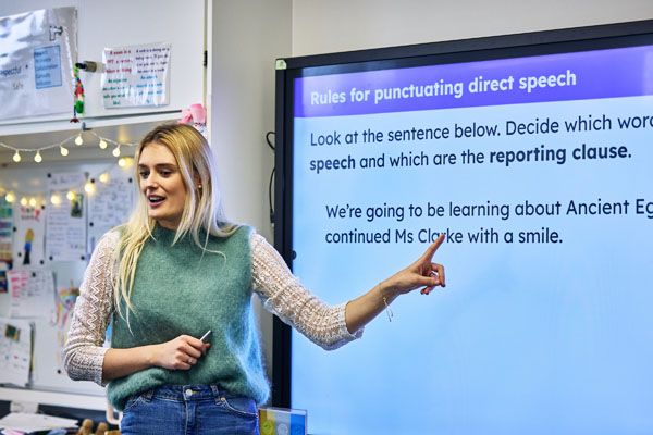 A primary teacher stood at the front of a class pointing to a smart board