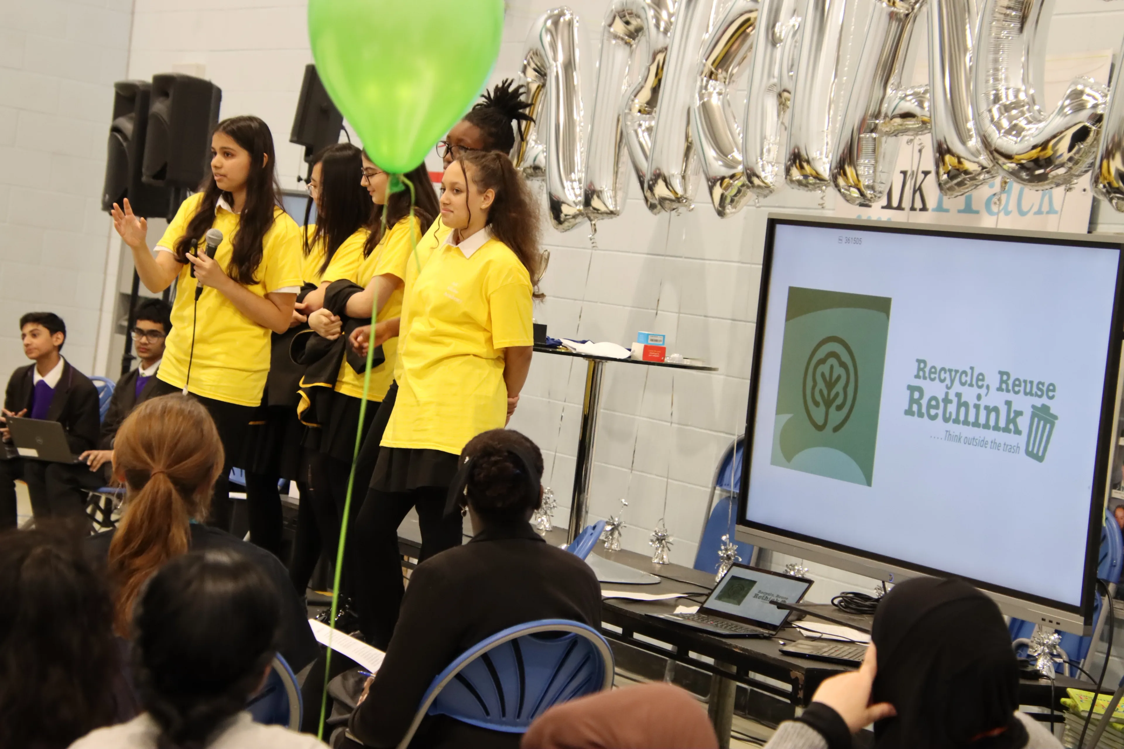 A group of pupils standing in front of a room, speaking and delivering their Arkhack pitch. There is also a large television at the front of the room displaying a slide deck of their pitch.