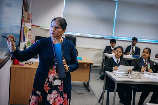 A teacher at the front of a class interacting with a smart board with pupils sat at desks looking towards the smart board.