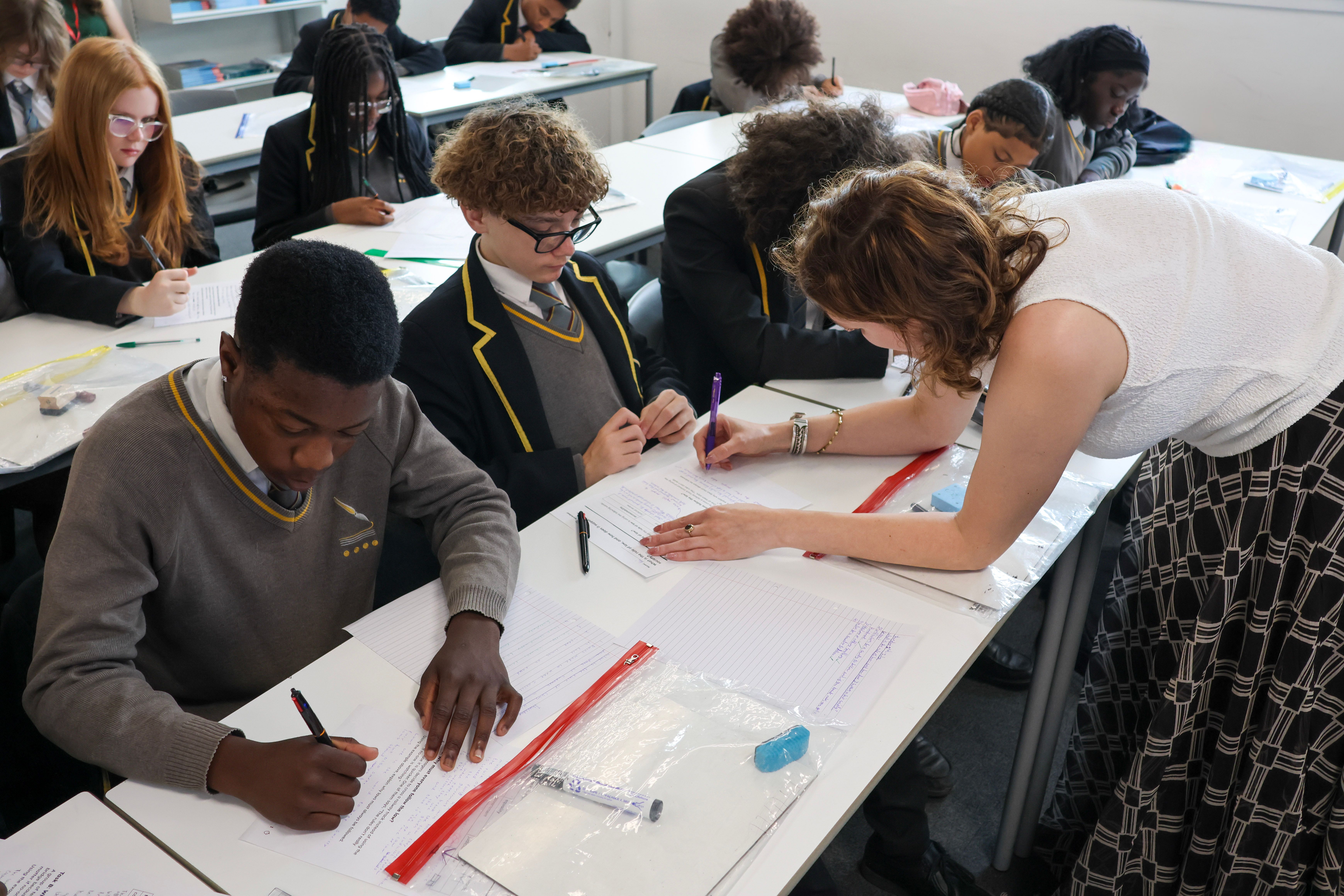 A teacher supports a pupil with a worksheet while other pupils work at their desks in a classroom.