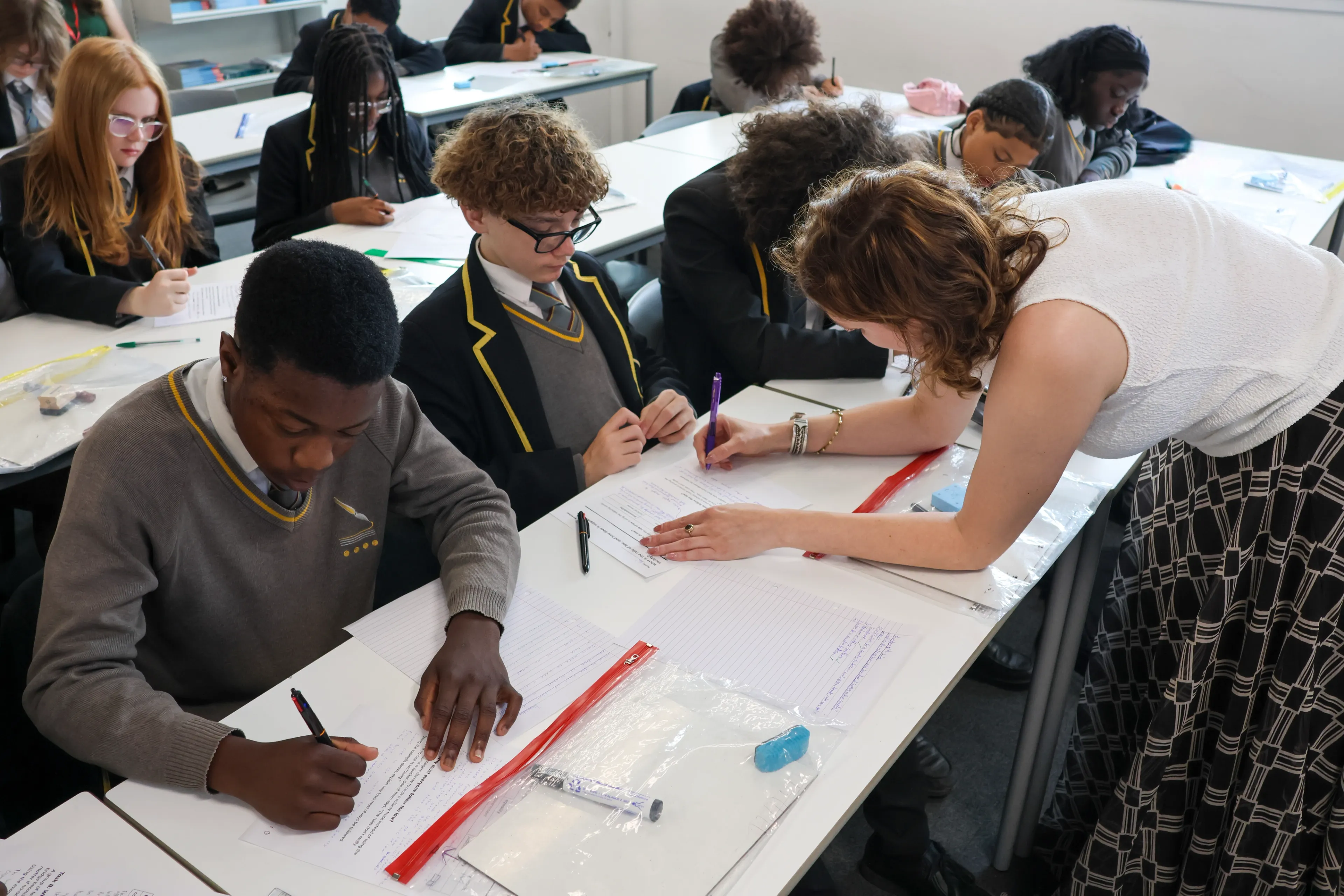 A teacher supports a pupil with a worksheet while other pupils work at their desks in a classroom.