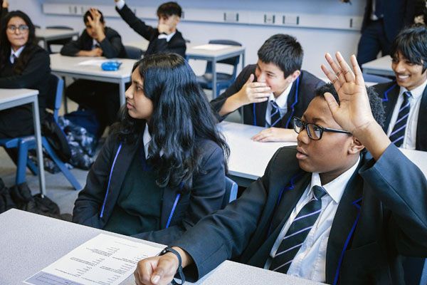 Pupils sat in a classroom at desks, some pupils are raising their hands