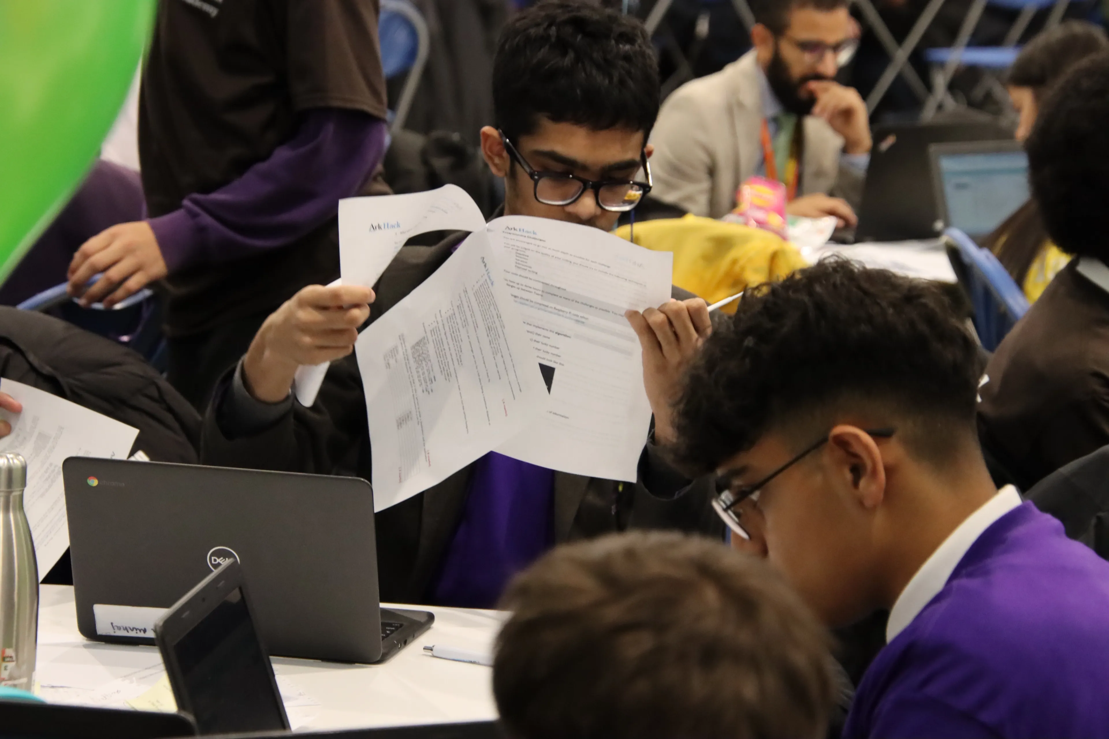 A group of pupils sitting at desks concentrating hard. They are surrounded by paper work and laptops and are working on their ArkHack projects
