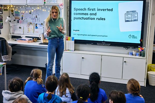 A primary school teacher is standing in front of a whiteboard that reads 'speech first: inverted commas and punctuation rules. English, Oak National Academy', teaching a class of primary school children sitting on the floor. 